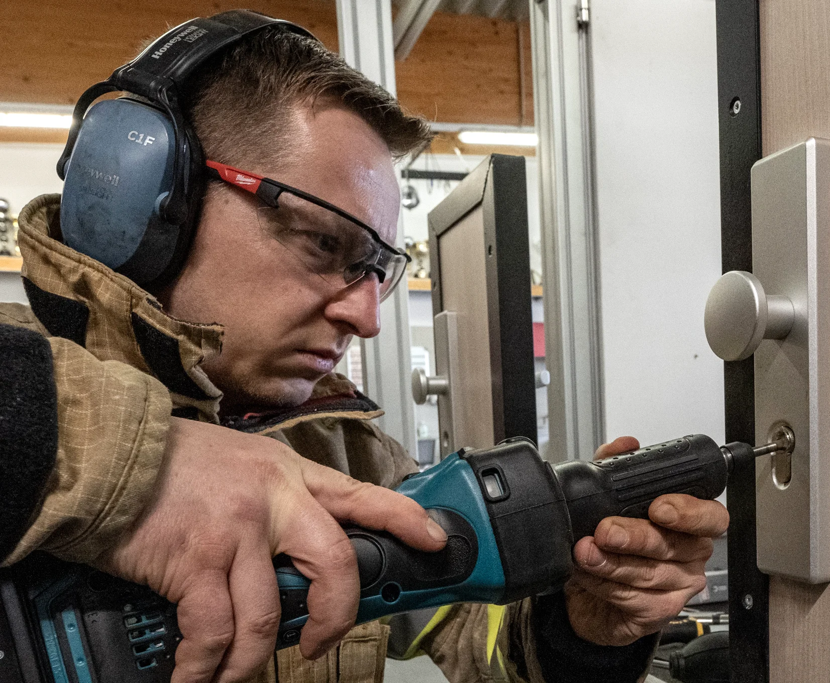 Fachkraft mit Schutzbrille und Gehörschutz bohrt in einer Werkstatt einen Türzylinder an einer Übungstür.