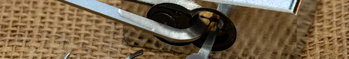 Nahaufnahme eines Profilzylinders, der mit professionellen Lockpicking-Werkzeugen geöffnet wird. Ein Spanner und ein Dietrich befinden sich im Schlüsselloch.