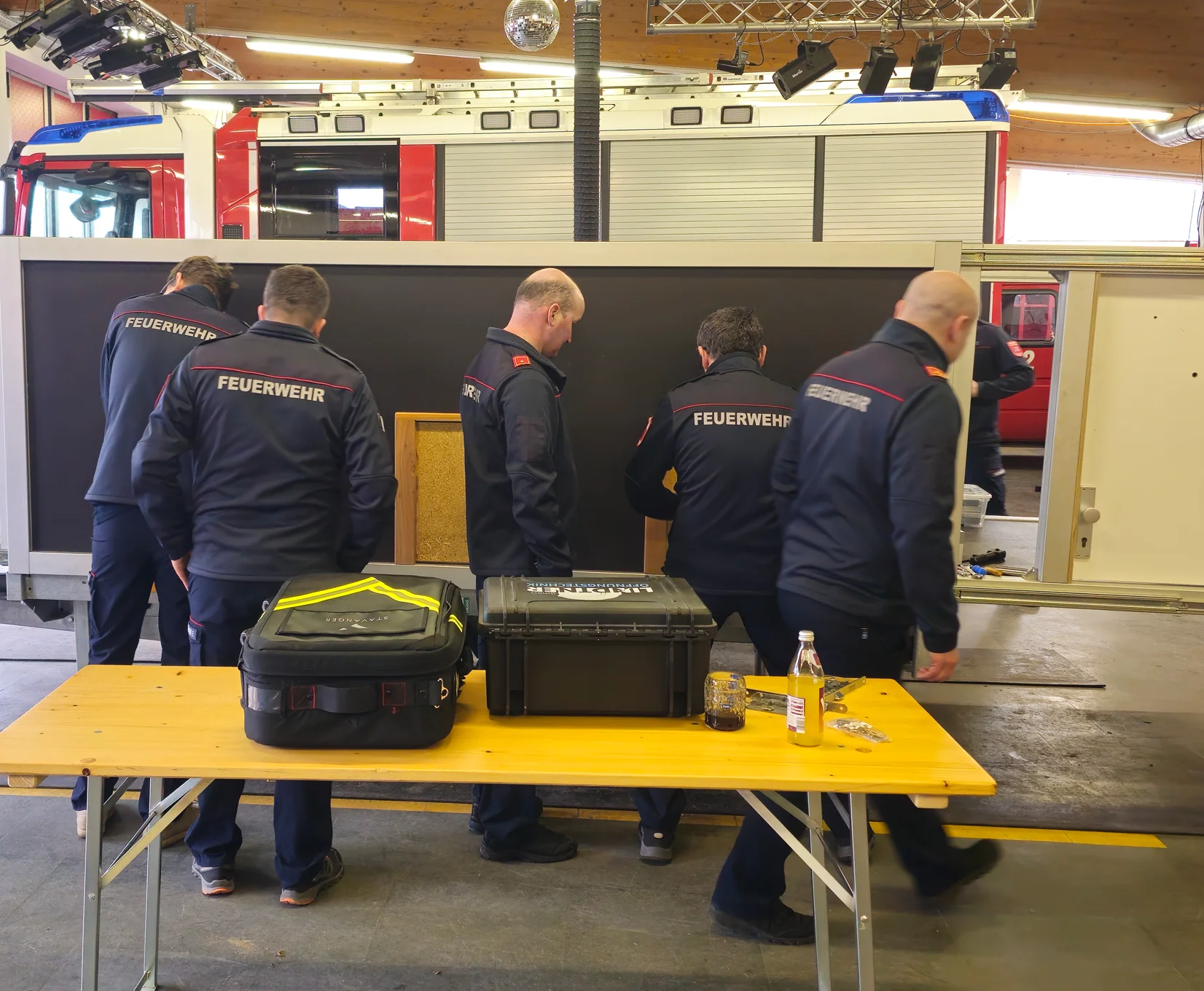 Feuerwehrkräfte bei einer praktischen Schulung an einer Übungswand, davor ein Tisch mit Werkzeugkoffern und Ausrüstung in einer Fahrzeughalle.